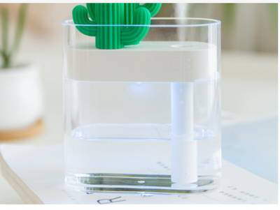 Transparent portable humidifier with a green cactus design on top, emitting mist on a wooden desk next to a laptop. - Image 9
