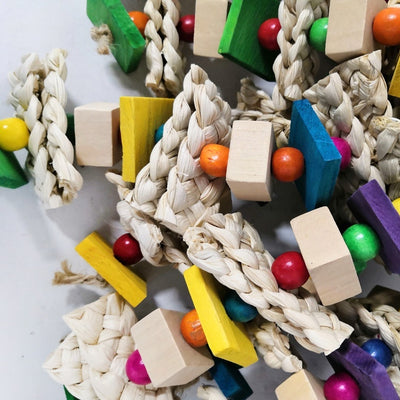 Colorful bird toy made of natural wood blocks, beads, and corn husk strips, with a red and green macaw.