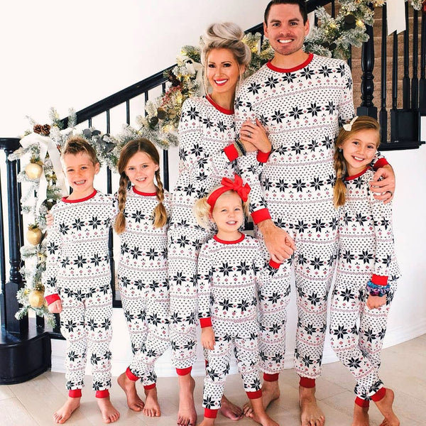 Family of six wearing matching white Christmas pajamas with black snowflake and red trim, posing by a decorated staircase.