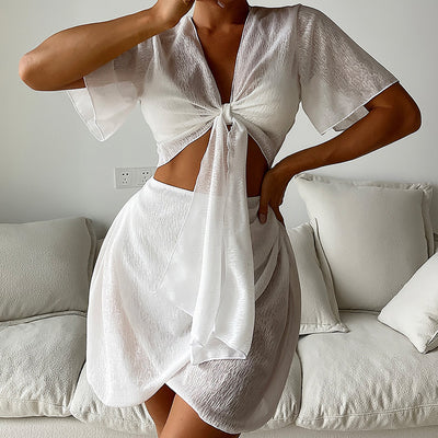 White two-piece swimwear set with a tie-front crop top and a high-waisted skirt with an asymmetrical ruffled hem.