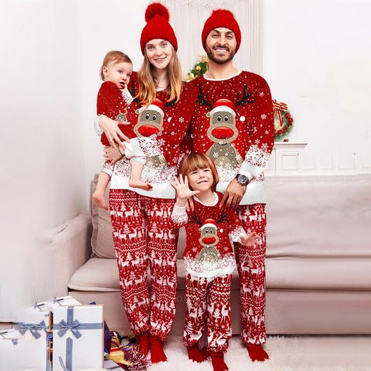 Family of four wearing matching red Christmas pajamas with reindeer and snowflake print, plus red pom-pom beanies.