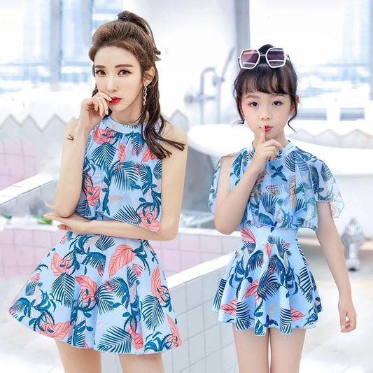 Mother and daughter matching swimsuits with blue and pink tropical leaf print. Adult halter top and skirt, child's ruffle top and skirt.