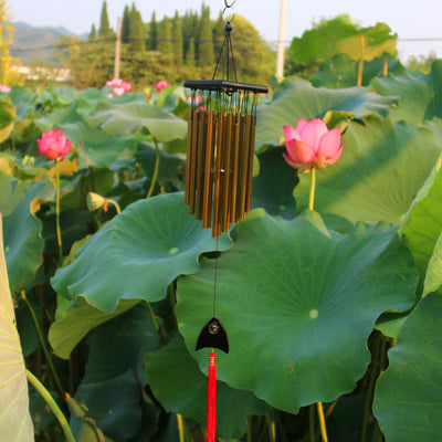 Bronze wind chime with multiple metal tubes hanging from a dark rectangular top, set against green lotus leaves. - Image 9