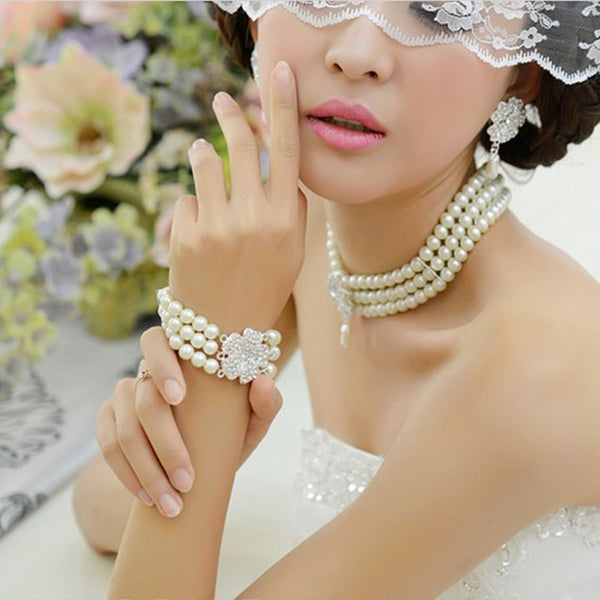 Three-piece pearl jewelry set with multi-strand necklace, bracelet, and drop earrings, all featuring sparkling rhinestone flower accents.