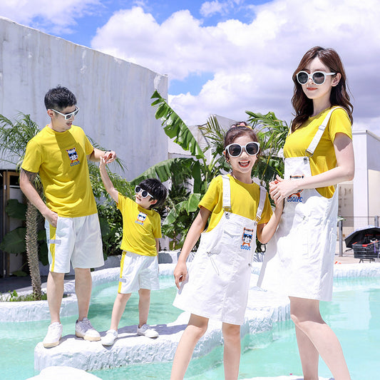 Family of three in matching yellow t-shirts and white outfits by a pool. Father and son wear shorts, mother and daughter wear overall dresses.