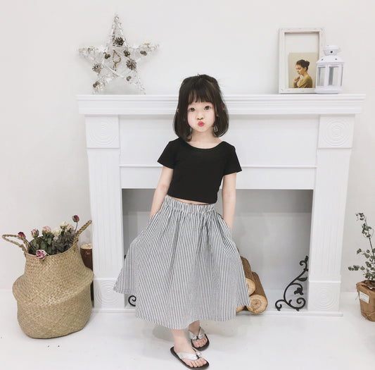 Young girl wearing a black short-sleeve crop top and a grey and white striped midi skirt with flip-flops.