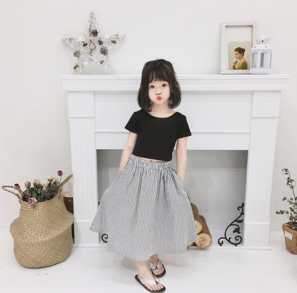 Young girl wearing a black short-sleeve crop top and a grey and white striped midi skirt with flip-flops.