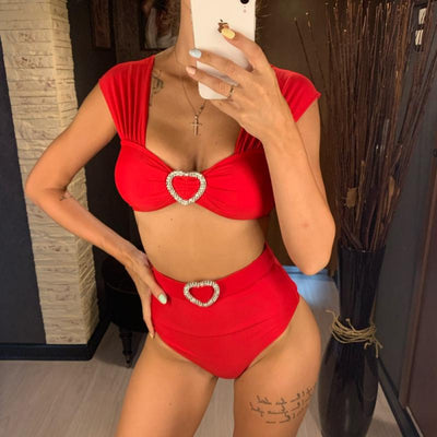 Red high-waisted two-piece bikini with a bandeau top and heart-shaped rhinestone buckles, displayed on a wooden surface.