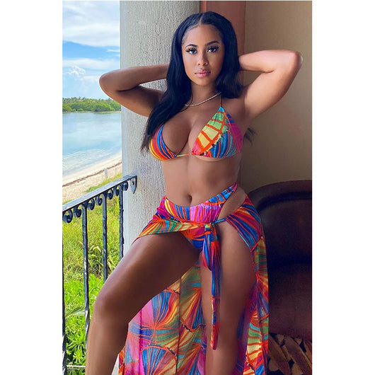 Woman wearing a vibrant multi-color abstract print three-piece bikini set with a matching sheer sarong on a balcony.