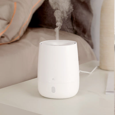 White minimalist aroma diffuser emitting mist, with a single button, on a white nightstand next to a bed. - Image 3