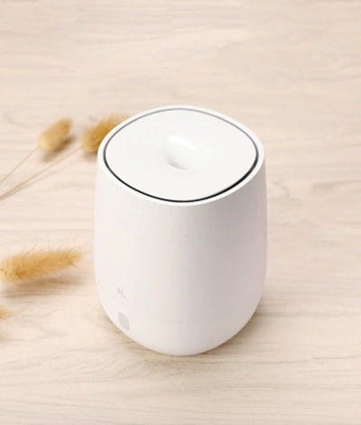 White minimalist aroma diffuser emitting mist, with a single button, on a white nightstand next to a bed. - Image 4