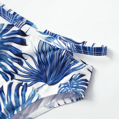 Family matching swimsuits with white and blue tropical leaf print, including men's shorts, women's and girl's ruffled one-shoulder bikinis. - Image 5