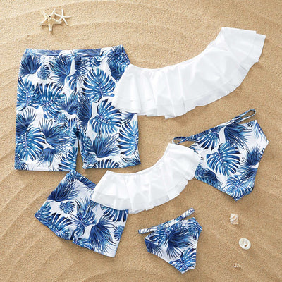 Family matching swimsuits with white and blue tropical leaf print, including men's shorts, women's and girl's ruffled one-shoulder bikinis.