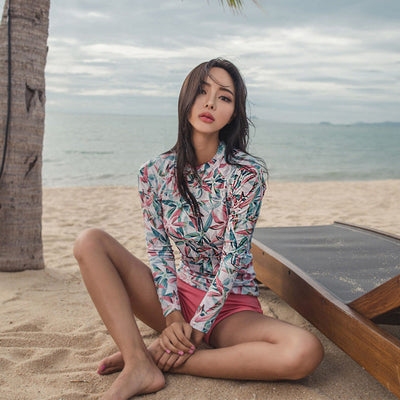 Woman in a long-sleeve tropical print rash guard and pink bikini bottoms sitting on a sandy beach. - Image 6