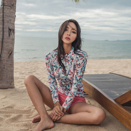 Woman in a long-sleeve tropical print rash guard and pink bikini bottoms sitting on a sandy beach. - Image 6
