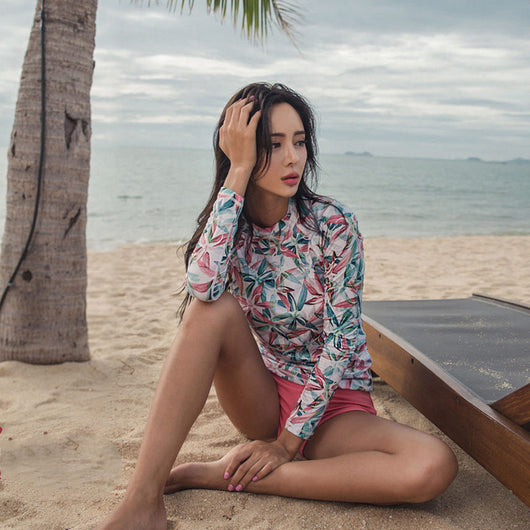 Woman in a long-sleeve tropical print rash guard and pink bikini bottoms sitting on a sandy beach. - Image 5