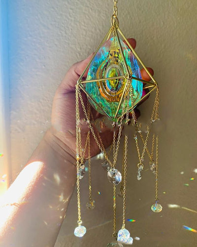 Geometric iridescent suncatcher with a gold frame and dangling crystal pendants, held in a hand, creating rainbow light.