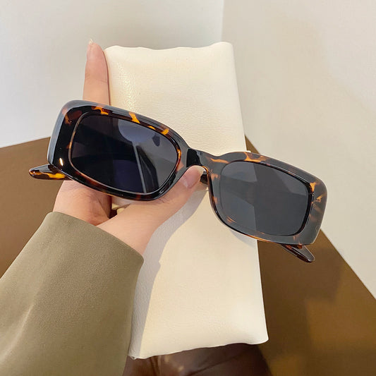 Black rectangular retro sunglasses with dark lenses held by a hand over a cream display pillow.