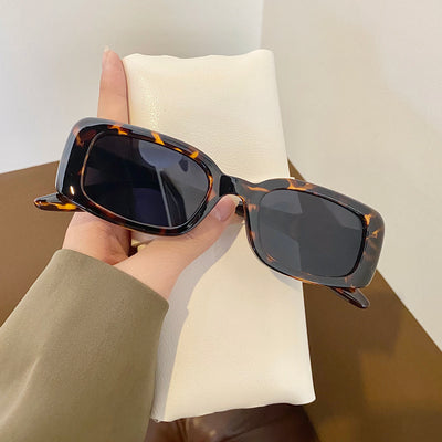 Black rectangular retro sunglasses with dark lenses held by a hand over a cream display pillow.