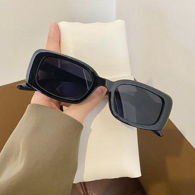 Black rectangular retro sunglasses with dark lenses held by a hand over a cream display pillow. - Image 3
