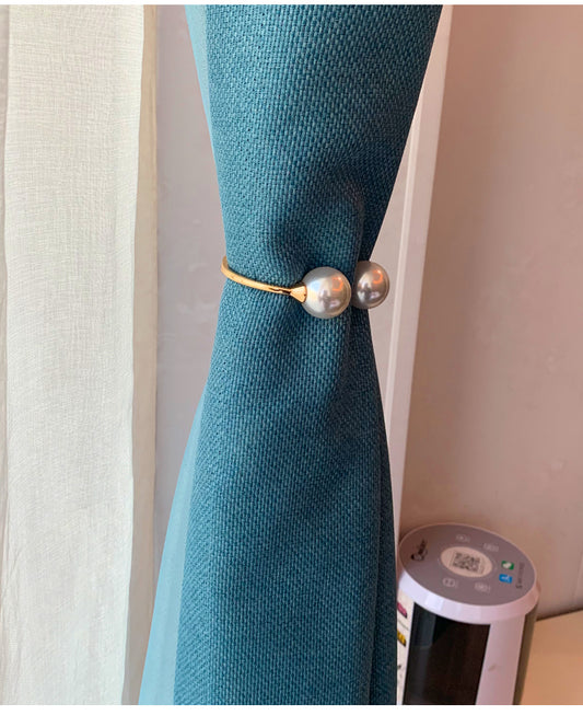 Gold alloy curtain tie-back with two large white faux pearls holding back a teal woven curtain.