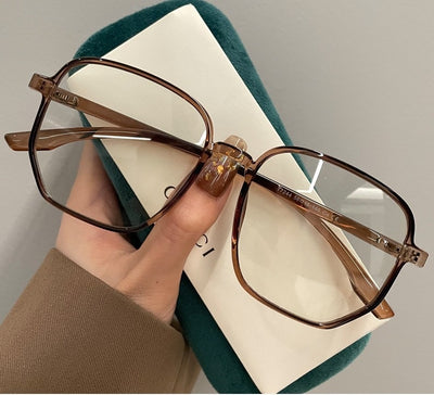 Mocha brown irregular frame glasses with clear lenses, held over a dark green velvet glasses case. - Image 12