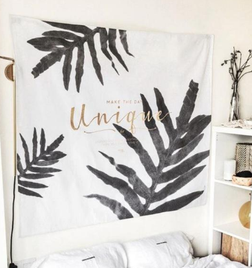 White polyester tapestry with black tropical leaf print and gold "Unique" script, hanging above a bed.