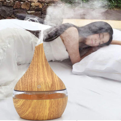 Dark wood grain LED essential oil diffuser with a blue light band and touch switch, shown with a hand operating it. Includes a bundle of 10 white filter sticks.