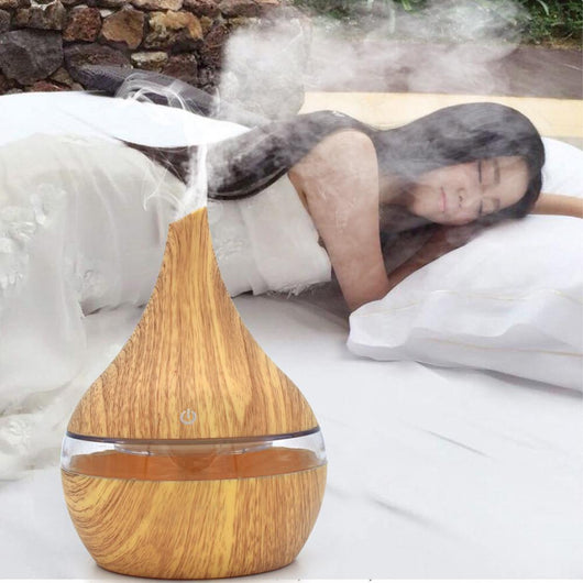 Dark wood grain LED essential oil diffuser with a blue light band and touch switch, shown with a hand operating it. Includes a bundle of 10 white filter sticks.
