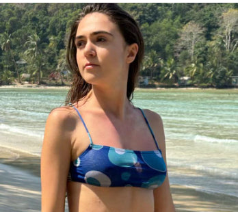 Woman in a blue banded bikini with a circular pattern standing on a sandy beach with clear water and green hills. - Image 9
