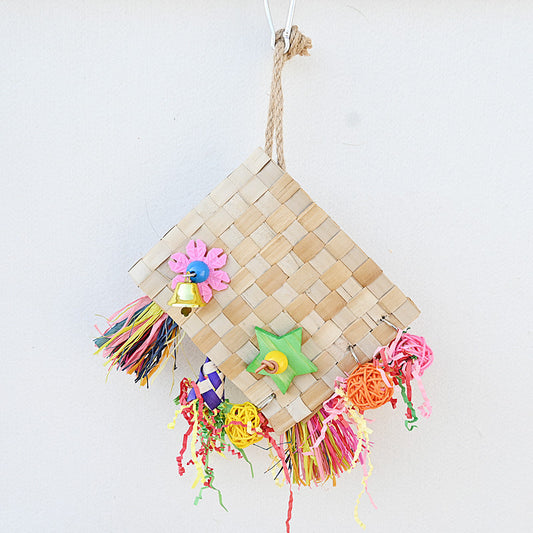 Colorful parrot foraging toy with woven palm leaf, a bell, wooden star, and shredded paper, hanging against a white wall.