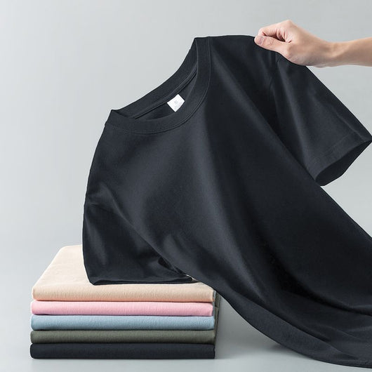Light gray short-sleeve t-shirt with a round neck, held up to show its smooth fabric. A stack of folded t-shirts in various colors is visible in the background. - Image 5