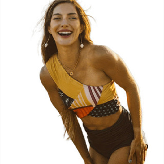 Smiling woman in a one-shoulder, color-block bikini top and high-waisted bottom on a sandy beach. - Image 4