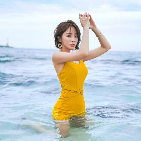 Woman in a mustard yellow one-piece swimsuit with a scalloped waist and short bottoms, standing in the ocean.
