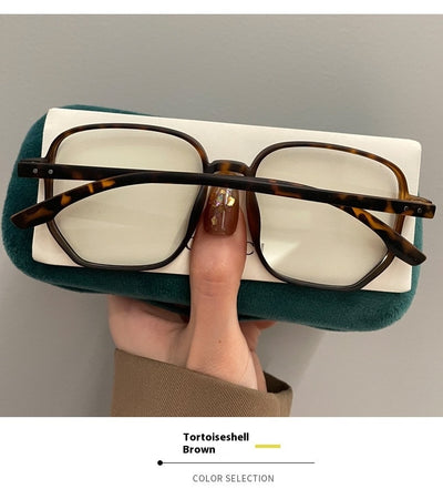 Mocha brown irregular frame glasses with clear lenses, held over a dark green velvet glasses case. - Image 16