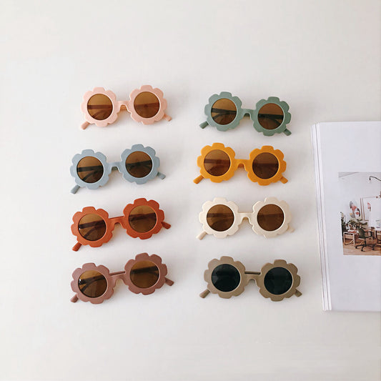 Cream flower-shaped sunglasses for babies and toddlers with dark lenses, displayed on crumpled grey paper.