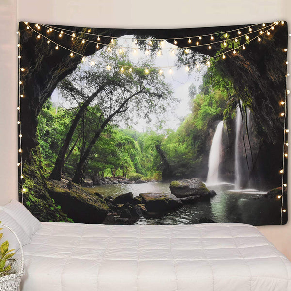 Cave waterfall print tapestry with lush green trees and dark rocks, adorned with warm string lights above a white bed.