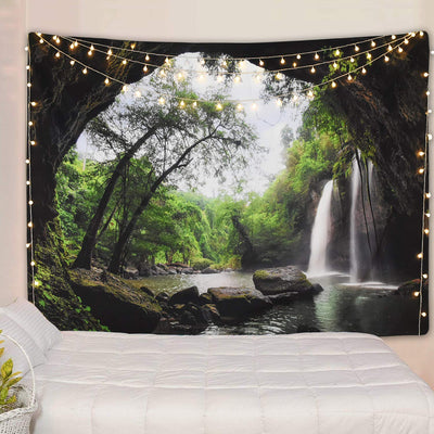 Cave waterfall print tapestry with lush green trees and dark rocks, adorned with warm string lights above a white bed.