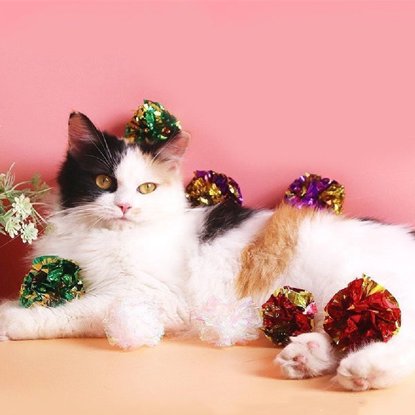 Five iridescent crinkle paper balls, likely cat toys, arranged in a circle on a peach background.