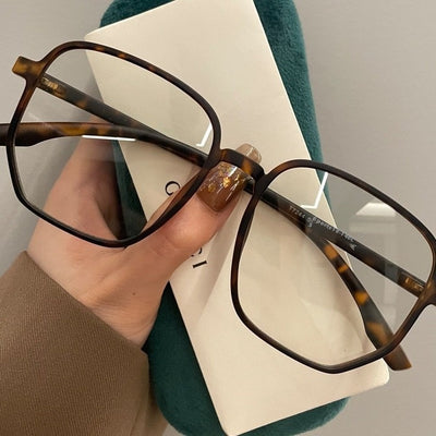 Mocha brown irregular frame glasses with clear lenses, held over a dark green velvet glasses case.