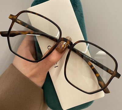 Mocha brown irregular frame glasses with clear lenses, held over a dark green velvet glasses case. - Image 14
