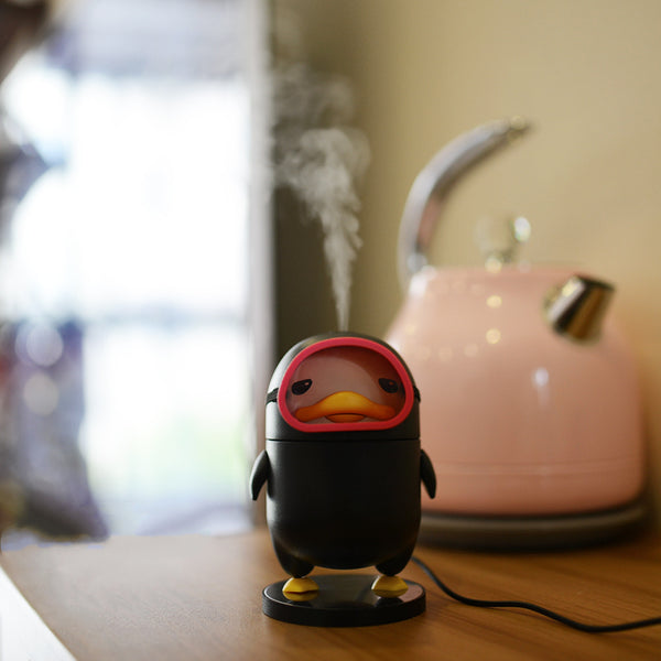 Black diving duck spray humidifier with red goggles and yellow feet, standing on a dark base on a table.