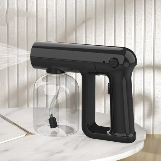 Black wireless alcohol blue light disinfection spray gun with clear reservoir, actively misting, on a marble surface.