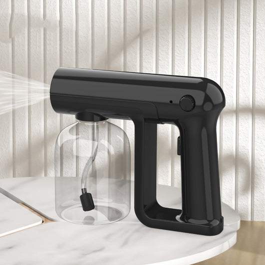 Black wireless alcohol blue light disinfection spray gun with clear reservoir, actively misting, on a marble surface.