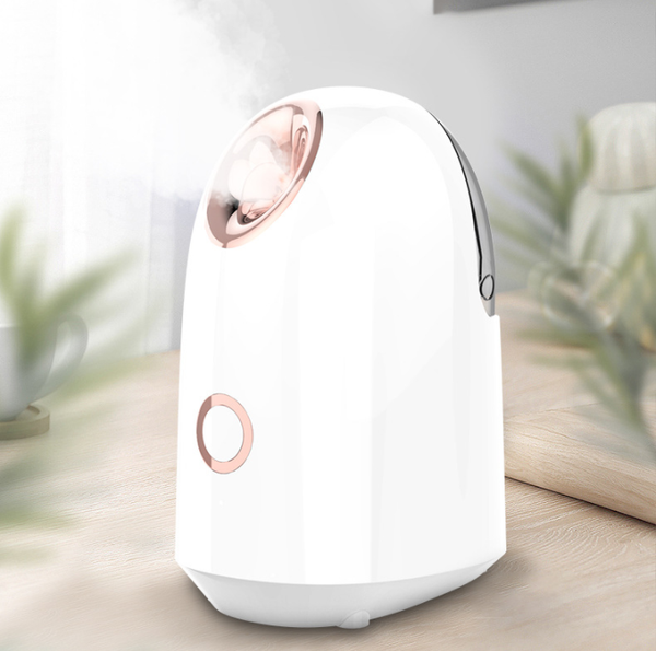 White and rose gold steam facial sprayer emitting warm mist, shown on a light wooden table.