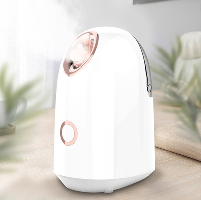 White and rose gold steam facial sprayer emitting warm mist, shown on a light wooden table.