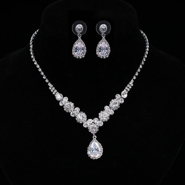Sparkling silver-toned necklace and matching teardrop earrings set with clear stones, displayed on a black stand.