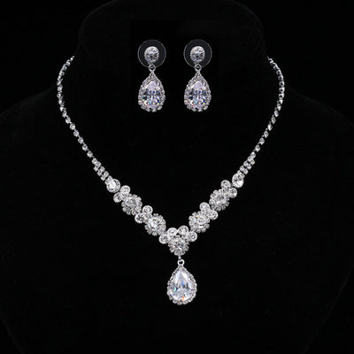 Sparkling silver-toned necklace and matching teardrop earrings set with clear stones, displayed on a black stand.