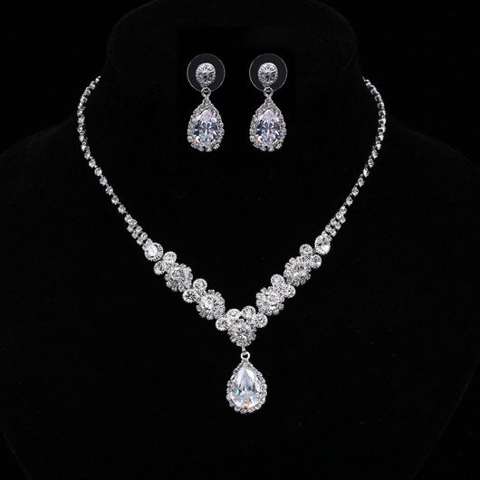 Sparkling silver-toned necklace and matching teardrop earrings set with clear stones, displayed on a black stand.