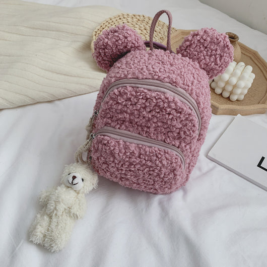 Pink fuzzy mini backpack with bear ears and a small white teddy bear keychain on a white bed.